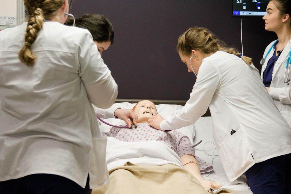护理系学生观看女护理系学生练习从躺在医院病床上的人体模型上取生命体征